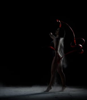 beautiful caucasian woman in a black bodysuit with a sports figure dancing with red ribbon on a black background, particles of flour fly in different directions