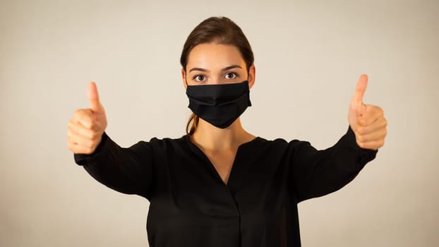 Positive woman wearing black face mask showing her thumbs up. Happy young Caucasian girl expressing encouragement and support with her fingers during medical crisis of coronavirus.