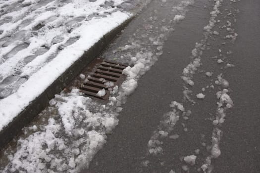 5 - Urban tarmac road and sidewalk covered in heavy snow that is beginning to thaw. Surface level storm drain deals with the resulting water.