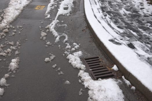 8 - Surface drain on the corner of a tarmac road, surrounded by melting snowfall. Winter environment in the UK, and water management infrastructure.