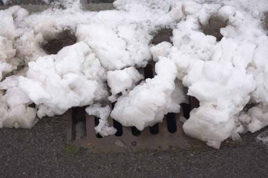 10 - Winter scene of a roadside drain covered in heavy chunks of white snow. The metal grill of the drain is nearly fully blocked by the snowfall.