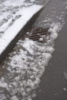 6 - Thawing snow flows into a metal curbside drain on the surface of a tarmac road. Track visible where a car has driven through the snow.