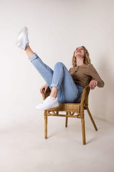 portrait of a beautiful young woman in jeans, shirt and sneakers.she put her feet up on the wicker rocking chair against the white brick wall.sitting on a chair.