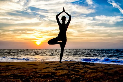 The figure of a girl on the background of the sea and sunset in a yoga pose on one leg.