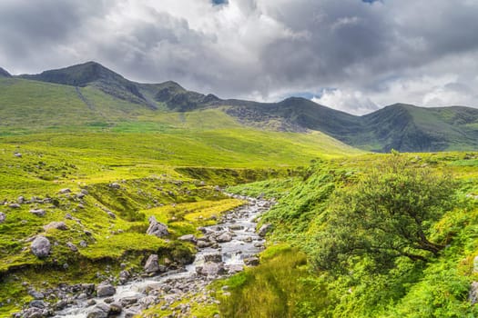 Winding mountain stream or river in Cronins Yard. Green fields in a valley with tall mountains covered in sunlight, Ring of Kerry, Ireland