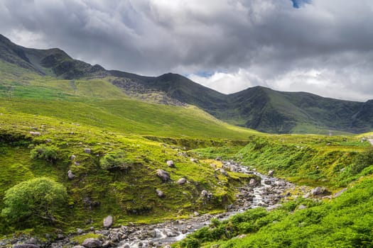 Winding mountain stream or river in Cronins Yard. Green fields in a valley with tall mountains covered in sunlight, Ring of Kerry, Ireland