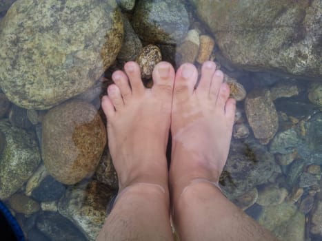 Top view of transparent water surface with human feet visible in it. Copy space for background and texture. Transparent water texture with view of stones under water.