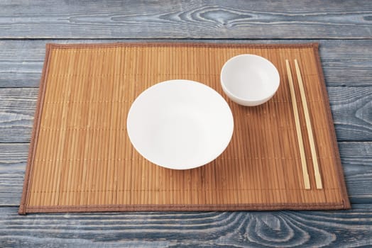 Two white empty bowl and two wooden chopsticks lie on a bamboo mat that sits on a wooden table made of textured boards.