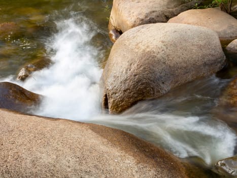 A long-exposure shot of a small creek flowing through a brown rock. And there are green trees as a backdrop. It is a specific focus.