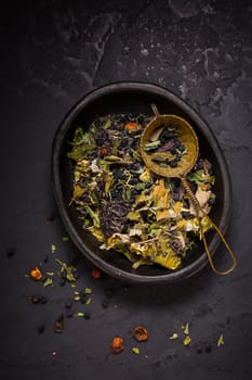 Mixture of teas, dried organic tea herbs and fruits in bowl with tea strainer on black background