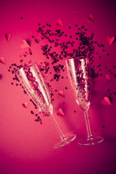 Two champagne glasses with splash of red heart shaped confetti on red background. Overhead view, copy space. Valentine's Day concept