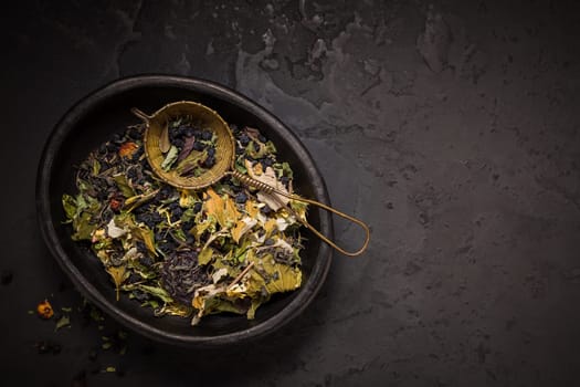 Mixture of teas, dried organic tea herbs and fruits in bowl with tea strainer on black background