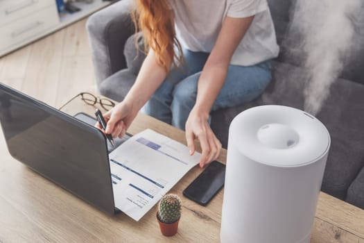 Woman freelancer uses a household humidifier in the workplace to maintain relative humidity and microclimate in the workplace of the home office with a laptop and documents.
