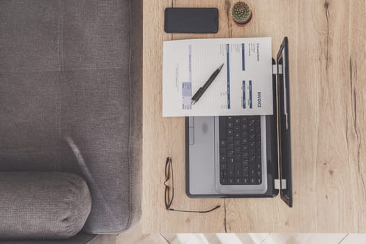 Flat lay of home office desk workspace. Laptop, invoice and eyeglasses on wooden table, sofa.