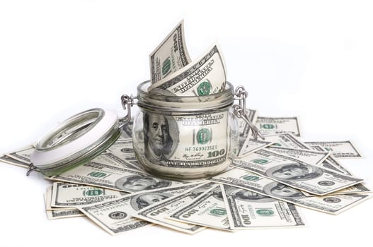 A glass jar filled with dollars among a hundred dollar bills on a white background.