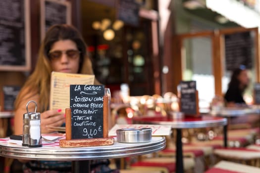 Beautiful young woman at a typical french restaurant. Placard says: Hot drink, orange juice, croissaint, pie and jam