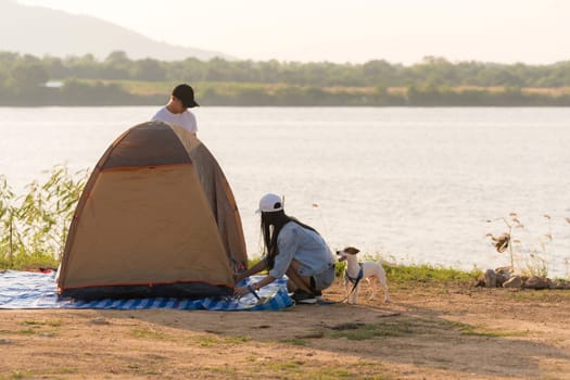 Young adult Asian couple pitch and set up tent for camping around the lake. Camping trip with dog outdoor activity campsite concept.