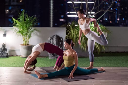 Group of asian women in sportswear pants practicing yoga during city lockdown in their apratment with distance in background of cityscape illumination night. Work out fitness healthy lifestyle concept