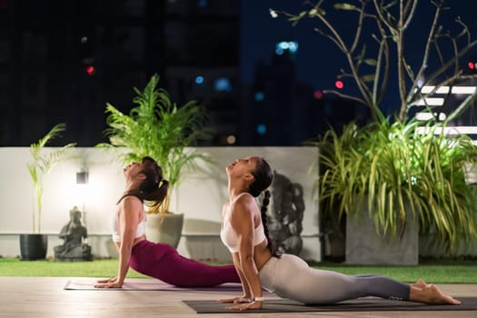 Two asian women in sportswear pants practicing yoga during city lockdown in their apratment with distance in background of cityscape illumination at night. Work out fitness healthy lifestyle concept.