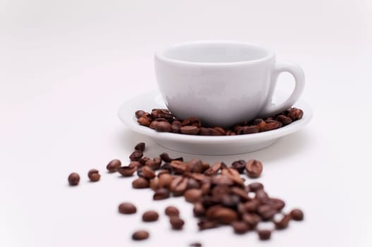 White cappuccino cup on saucer with roasted coffee beans on and in front of the saucer and white studio background.
