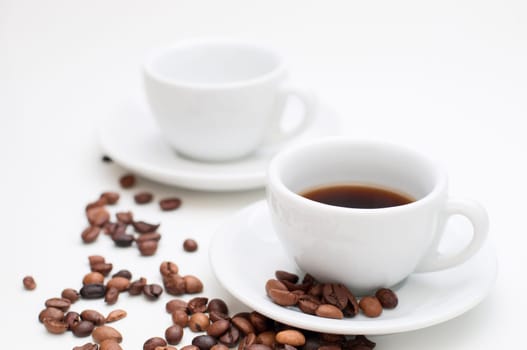 Two white cappuccino cups with saucers and scattered roasted coffee beans on white background. Focus is on the front cup filled with coffee beverage.