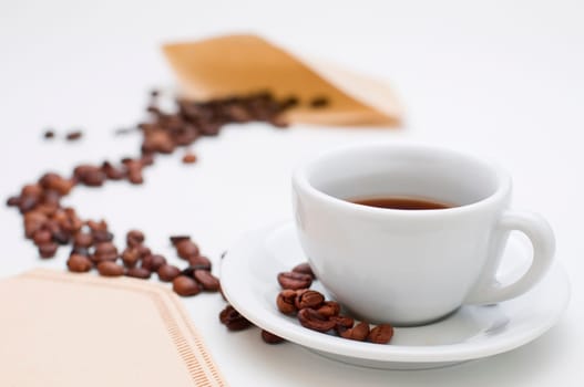 White half-filled cappuccino cup with saucer and individual roasted coffee beans in the sharp foreground and coffee beans arranged in an arch towards a filter bag in the blurred background on a white background.