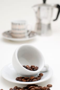 Tilted white espresso cup on white saucer with roasted coffee beans in, on and in front of the saucer in front of blurred cup with coffee pot on white background.