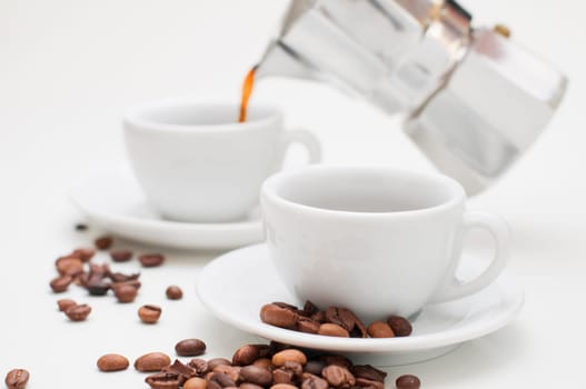 White cappuccino cup on white saucer with roasted coffee beans in focus in the foreground and a second cappuccino cup with pouring coffee pot in the blurred background.