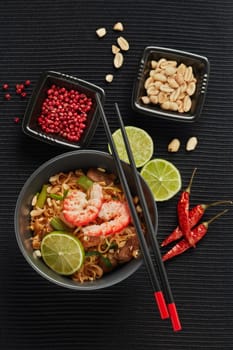 Pad Thai. Stir-fried rice noodles with chicken meat, shrimps,  scrambled egg, peanuts, vegetables and sauce in bowl on black background. Top view. Thai food. Setting table.