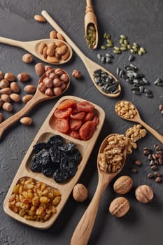 Various varieties of nuts, seeds and dried fruits lying in wooden spoones and dish on black slate background. Top view. Healthy food. Vegetarian nutrition