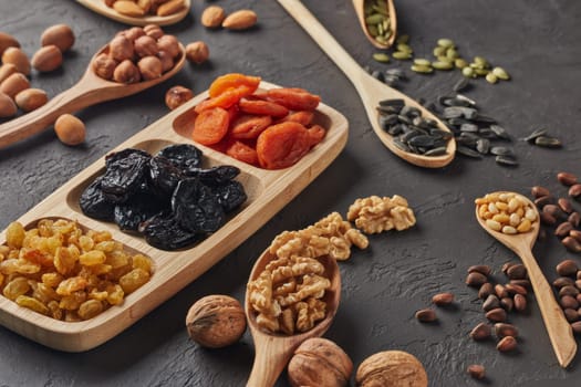 Various varieties of nuts, seeds and dried fruits lying in wooden spoones and dish on black slate background. Top view. Healthy food. Vegetarian nutrition