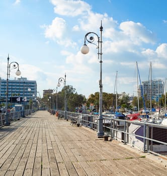 Wooden pier of marina with moored yachts and motorboats, hotels and apartments buildings in bright evening sunshnie, Larnaca, Cyprus
