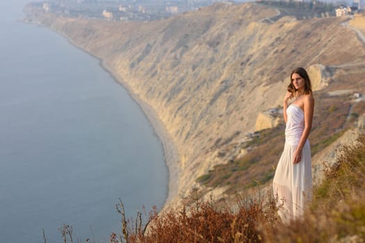 A beautiful slender girl in a white dress stands on a hill and enjoys a beautiful view of the sunset