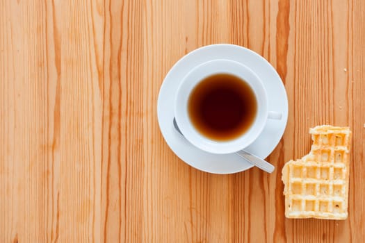 Wooden table with white cup with saucer and spoon as well as lying waffle with bite mark in top view.