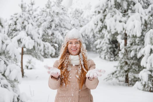Happy young redhead woman plays with snow in pine tree forest. Snow-covered fir trees on the background.