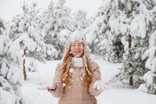 Happy young redhead woman plays with snow in pine tree forest. Snow-covered fir trees on the background.