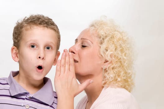 Head-and-shoulder view blonde mother whispers sideways into her 7-year-old son's ear with her hand held against a white background