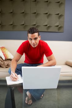 Portrait of a young man sitting in front of a laptop and writing on a spiral notebook