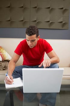 Young man sitting in front of a laptop and writing on a spiral notebook