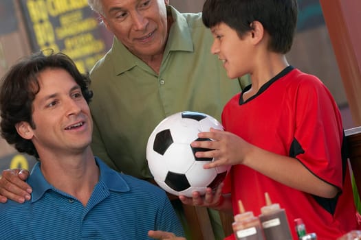 Close-up of a boy holding a soccer all with his father and grandfather looking at him