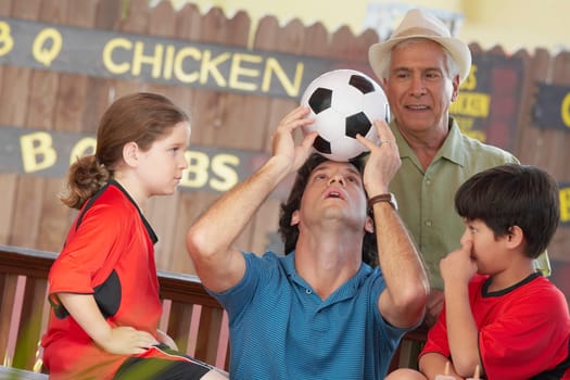 Mid adult man balancing a soccer ball on his forehead with his children and father looking at him
