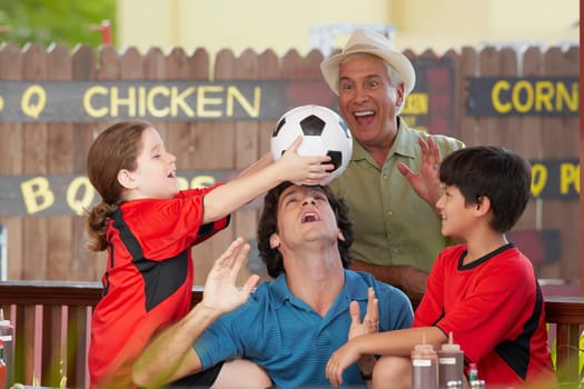 Side profile of a girl balancing a soccer all on her father's forehead with her brother and grandfather in a restaurant