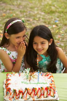 Girl sitting in front of a birthday cake and whispering to her friend