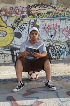 Portrait of a young man sitting on a soccer ball and holding a book