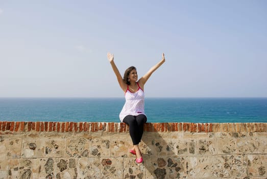Young woman sitting on the ledge with her arms raised and smiling