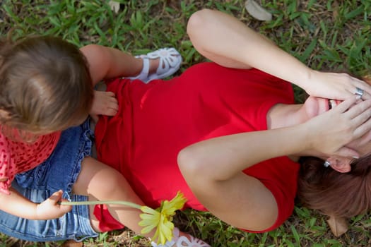 Young woman lying on grass and covering her face with her hands and her daughter sitting on her stomach