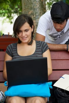 Young woman sitting on a bench and using a laptop with a young man leaning behind her