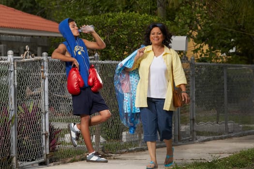 Mature woman walking with a teenage boy leaning on a chain-link fence and drinking water beside her