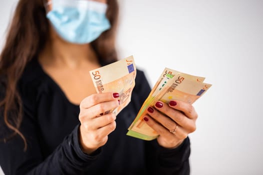 Young woman counting money and wearing blue surgical face mask during coronavirus pandemic with white background. Concept of economical problems of people caused by spreading illness.