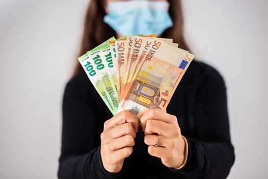 Hands holding euro cash during coronavirus pandemic. Concept of young Caucasian woman becoming rich. Human wearing face mask in studio and showing heap of money.